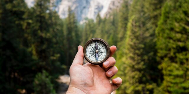 Person hält Kompass in die Luft. Im Hintergrund hat es Bäume und Berge.