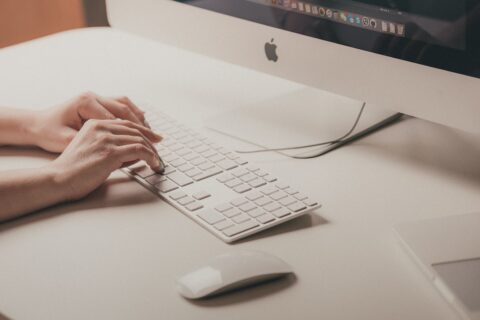 Person hält ihre Hände über der Tastatur eines Apple Computers. Auf dem Schreibtisch isch ausserdem eine Computermaus zu sehen.