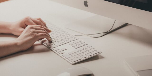 Person hält ihre Hände über der Tastatur eines Apple Computers. Auf dem Schreibtisch isch ausserdem eine Computermaus zu sehen.