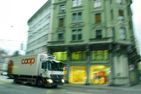 Verschwommener Coop Lieferwagen, der durch von links kommt und vor einem grossen Haus mit vielen Fenstern durchfährt. Zuunterst brennt Licht in den Schaufenstern.
