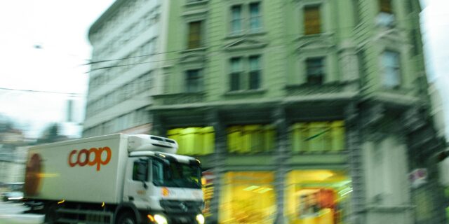 Verschwommener Coop Lieferwagen, der durch von links kommt und vor einem grossen Haus mit vielen Fenstern durchfährt. Zuunterst brennt Licht in den Schaufenstern.