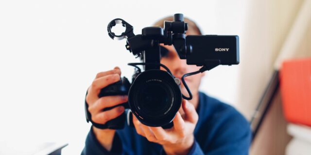 Person hält Kamera in der Hand und hat das Objektiv auf den Bildbetrachter gerichtet. Im Hintergrund ist verschwommen ein Raum zu sehen.