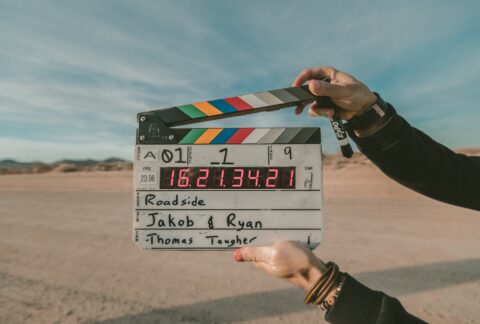 Eine Person hält eine Filmklappe mit einigen technischen Angaben vor die Kamera, Im Hintergrund sieht man eine Wüste mit einigen Hügeln.