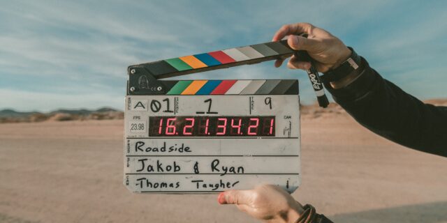 Eine Person hält eine Filmklappe mit einigen technischen Angaben vor die Kamera, Im Hintergrund sieht man eine Wüste mit einigen Hügeln.