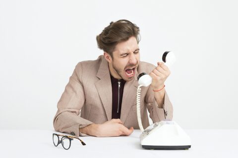 Mann hält den Hörer eines altenKabeltelefons in der Hand und brüllt hinein. Der gut gepflegte Mann trägt einen Anzug, seine Brille liegt vor ihm auf dem Tisch.