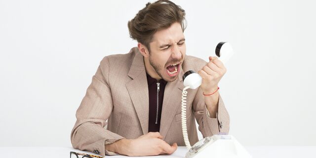 Mann hält den Hörer eines altenKabeltelefons in der Hand und brüllt hinein. Der gut gepflegte Mann trägt einen Anzug, seine Brille liegt vor ihm auf dem Tisch.