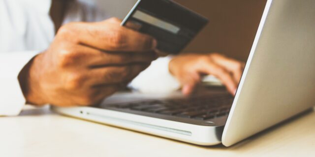 Person hält in der rechten Hand eine Kreditkarte, die linke Hand hält er über der Tastatur eines Laptops, der vor ihm auf einem Tisch steht.