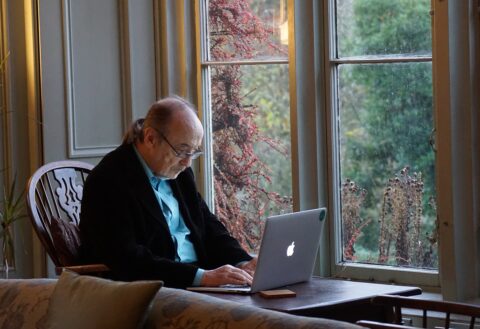 Älterer Herr sitzt in einem Sessel am Fenster und blickt auf einen Laptop, der vor ihm auf einem Tisch steht.