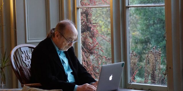 Älterer Herr sitzt in einem Sessel am Fenster und blickt auf einen Laptop, der vor ihm auf einem Tisch steht.