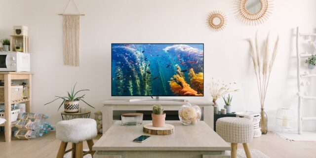 Ein Wohnzimmer mit einem Fernseher an der Wand. Der Bildschirm zeigt Unterwasseraufnahmen. Im Wohnzimmer stehen ein Tisch, Stühle und einige weitere Möbel.