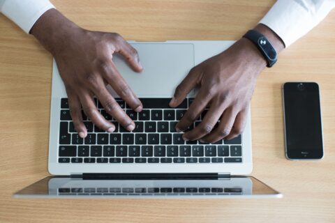 Eine Person sitzt an einem Schreibtisch und hält ihre Hände über der Laptoptastatur eines Macs.