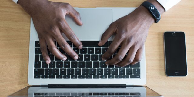 Eine Person sitzt an einem Schreibtisch und hält ihre Hände über der Laptoptastatur eines Macs.
