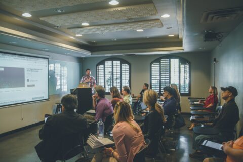 Seminarraum, ein Mann steht vorne und spricht. Im Raum sitzen etwa 15 Personen, die zuhören. An der vorderen Wand läuft eine PowerPoint Präsentation.