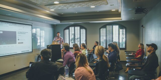 Seminarraum, ein Mann steht vorne und spricht. Im Raum sitzen etwa 15 Personen, die zuhören. An der vorderen Wand läuft eine PowerPoint Präsentation.