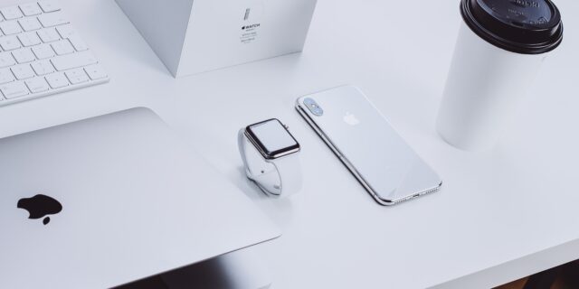 Auf einem Tisch liegen verschiedene Apple Produkte: ein MacBook, eine Apple Watch, ein iPhone, eine Bos und eine Tastatur. Ausserdem steht ein wegwerfbarer Kaffeebehälter auf dem Tisch.