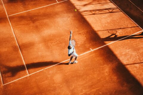Roter Tennisplatz von oben, ein Spieler hat einen Tennisball hochgeworfen und mit dem Schläger zum Schlag ausgeholt.