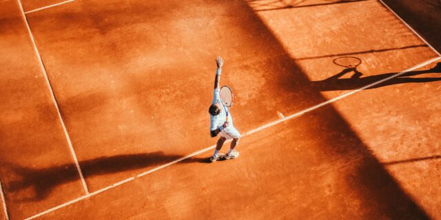 Roter Tennisplatz von oben, ein Spieler hat einen Tennisball hochgeworfen und mit dem Schläger zum Schlag ausgeholt.