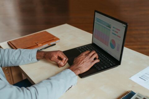 Eine Person sitzt an einem Schreibtisch und hät ihre Hände über einer Laptoptastatur. Auf dem Tisch liegen auch ein Heft und einige Blätter.
