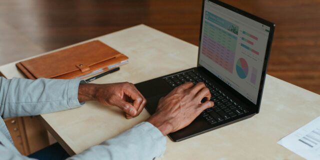 Eine Person sitzt an einem Schreibtisch und hät ihre Hände über einer Laptoptastatur. Auf dem Tisch liegen auch ein Heft und einige Blätter.