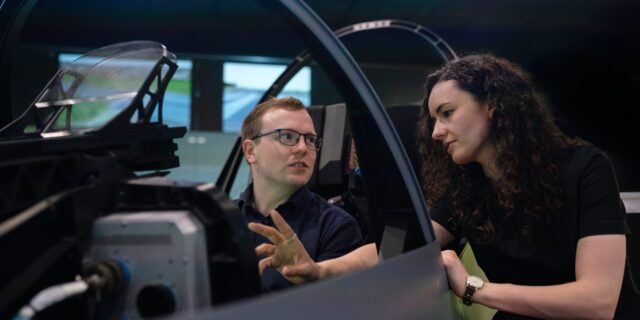 Zwei junge Menschen sitzen im Flugsimulator. Eine Frau erklärt einem Mann etwas.