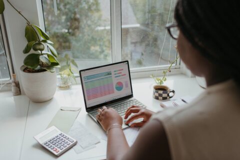 Eine Frau sitzt an einem Tisch und arbeitet an einem Windows Notebook. Auf dem Tischstehen einige Pflanzen und andere Arbeitsutensilien. Hinter dem Tisch ist ein Fenster.