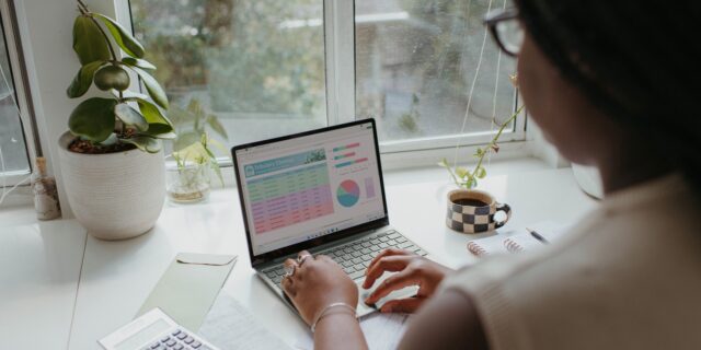 Eine Frau sitzt an einem Tisch und arbeitet an einem Windows Notebook. Auf dem Tischstehen einige Pflanzen und andere Arbeitsutensilien. Hinter dem Tisch ist ein Fenster.