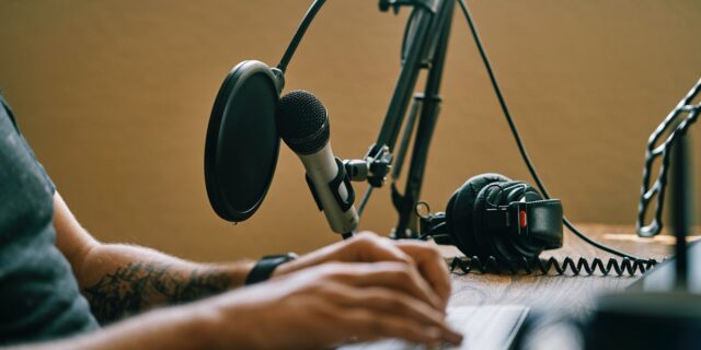 Eine Person mit tätowierten Armen sitzt an einem Schreibtisch und hat seine Hände auf einer Tastatur. Vor ihm steht ein Mikrophon, es liegen Kopfhörer auf dem Tisch.