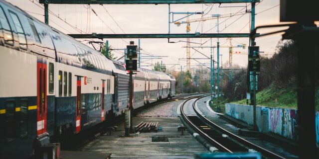 Zwei Gleise, die eine Kurve nach rechts machen. Auf dem linken Gleis fährt ein Zug. Im Hintergrund sind Bäume und Baukrane zu sehen. Der Himmel ist bewölkt.