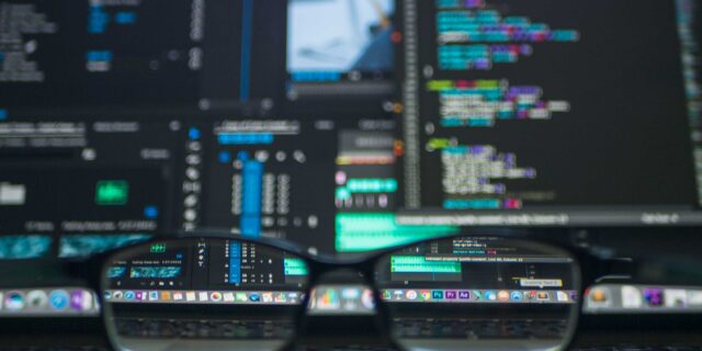 Verschwommener Computerbildschirm im Hintergrund, im Vordergrund liegt eine Brille, durch deren Gläser ein kleiner Teil des Bildschirms scharf wird. Es sind verschiedene geöffnete Programme erkennbar.
