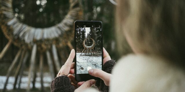 Frau hält Smartphone in den Händen und nimmt ein Foto auf von einem Bauwerk aus Holz am Waldrand.