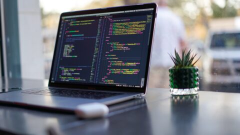 Ein Laptop auf einem Tisch vor einem Fenster. Auf dem Bildschirm ist HTML Auszeichnungssprache zu erkennen