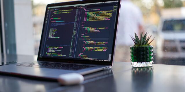 Ein Laptop auf einem Tisch vor einem Fenster. Auf dem Bildschirm ist HTML Auszeichnungssprache zu erkennen