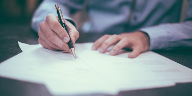 Ein Mann hält einen Stift in der Hand und schreibt etwas auf ein vor ihm auf einem Tisch liegendes Blatt.