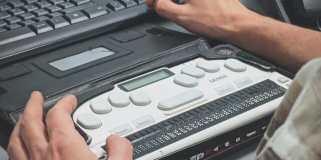 Blick über die Schulter einer Person, die vor einem Laptop mit Tastatur und Braillezeile sitzt