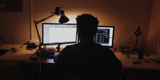 Person von hinten, die am Schreibtisch vor zwei Bildschirmen sitzt. Der Raum ist dunkel, eine Schreibtischlampe beleuchtet den Arbeitsplatz.