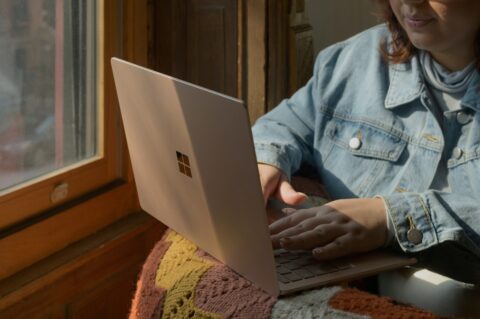 Junge Frau mit Microsoft Surface auf dem Schoss sitzt am Fenster.