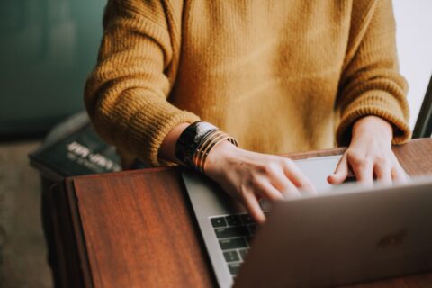 Person in gelbem Pullover sitzt am Schreibtisch am PC.