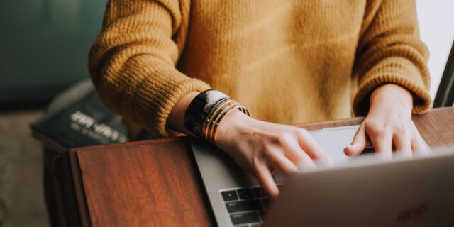 Person in gelbem Pullover sitzt am Schreibtisch am PC.
