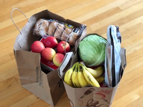 Zwei gefüllte Papiertragetaschen am Boden. Sie sind gefüllt mit Äpfeln, Brot und weiteren Lebensmitteln.