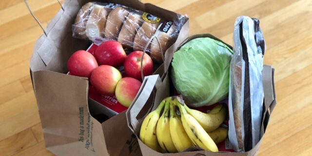 Zwei gefüllte Papiertragetaschen am Boden. Sie sind gefüllt mit Äpfeln, Brot und weiteren Lebensmitteln.