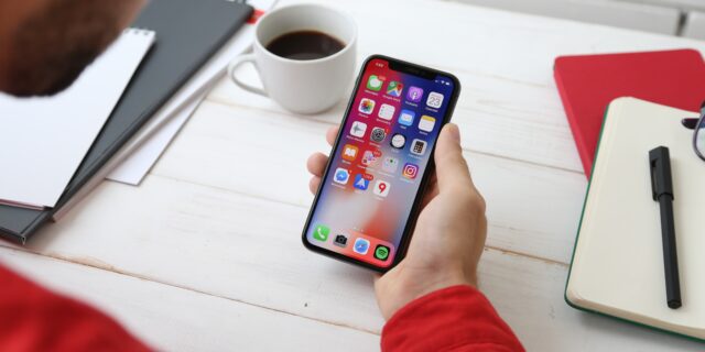Eine Person sitzt an ihrem Schreibtisch und hält ein Smartphone in der Hand. Auf dem Bildschirm ist der Homescreen mit vielen Apps zu sehen.