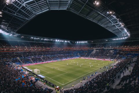 Gefülltes Fussballstadion bei Nacht. Die Spieler zweier Teams stehen verteilt auf dem beleuchteten Rasen