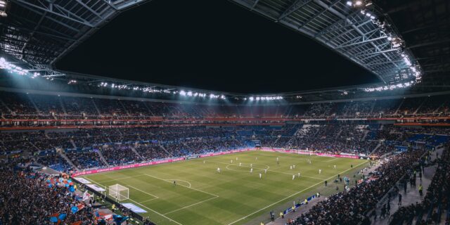 Gefülltes Fussballstadion bei Nacht. Die Spieler zweier Teams stehen verteilt auf dem beleuchteten Rasen