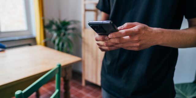 Mann in Wohnzimmer mit Smartphone in den Händen. Die Daumen sind auf dem Bildschirm.