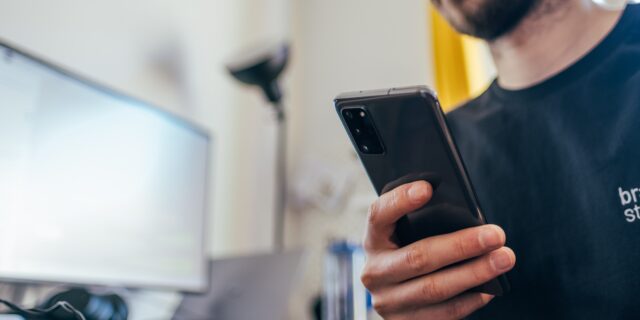 Mann sitzt am Schreibtisch und hält Smartphone in seiner rechten Hand.