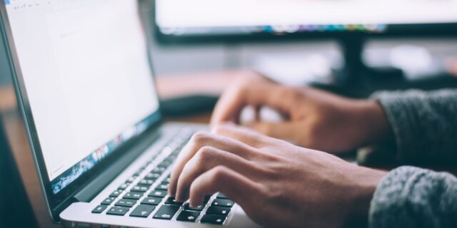 Hände einer Person über der Tastatur eines aufgeschlagenen Laptops. Der Bildschirm leuchtet.