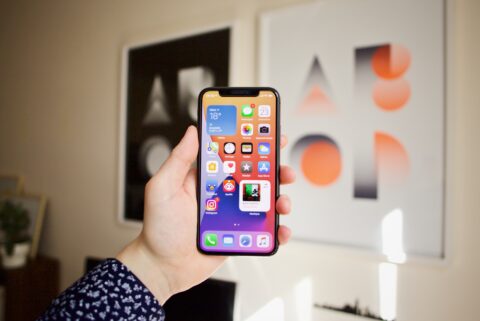 Eine Hand, die ein Apple Smartphone in der Hand hält. Im Hintergrund sieht man eine Wand mit zwei Bildern.