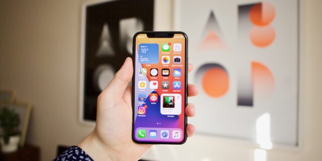 Eine Hand, die ein Apple Smartphone in der Hand hält. Im Hintergrund sieht man eine Wand mit zwei Bildern.