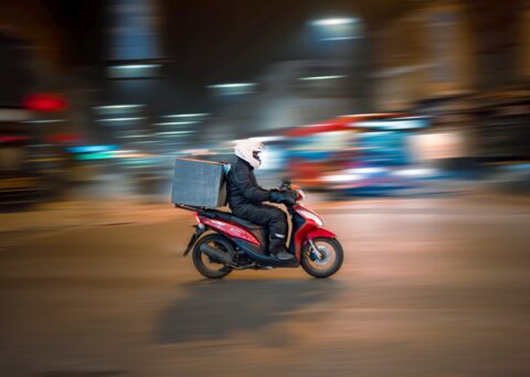Essenslieferant auf Motorrad unterwegs mit Kiste auf Gepäckträger. Der Hintergrund zeigt eine belebte Strasse bei Nacht und verschwommen.