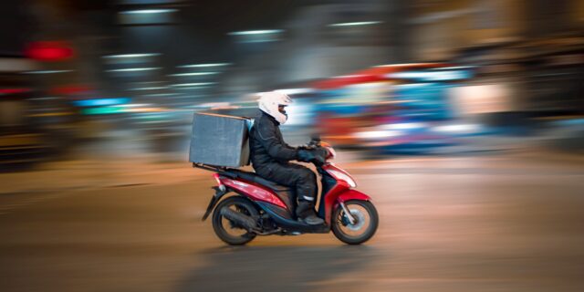 Essenslieferant auf Motorrad unterwegs mit Kiste auf Gepäckträger. Der Hintergrund zeigt eine belebte Strasse bei Nacht und verschwommen.